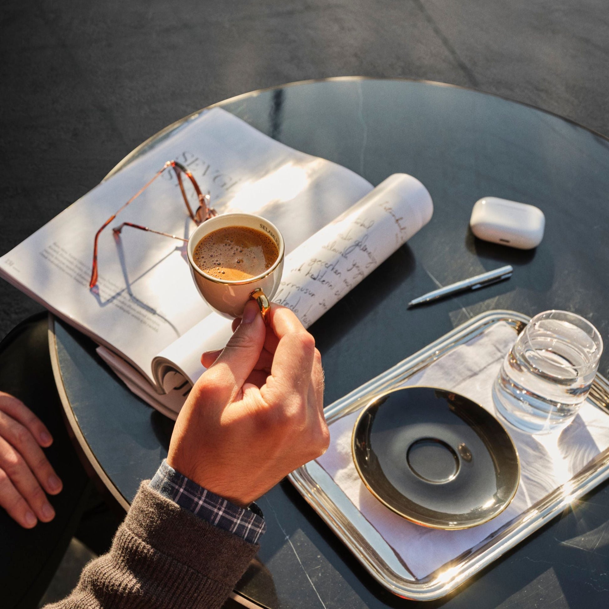 Person holding a small cup of coffee on a table with a book and glasses.